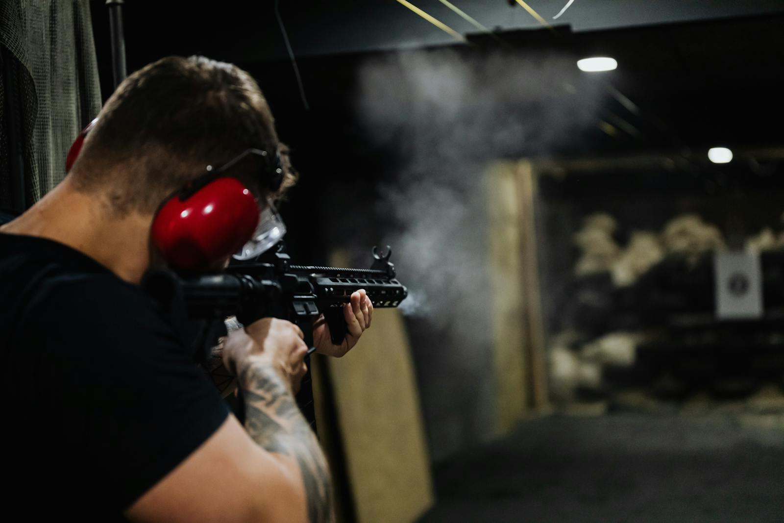 Adult man aiming a rifle at an indoor shooting range with smoke visible.